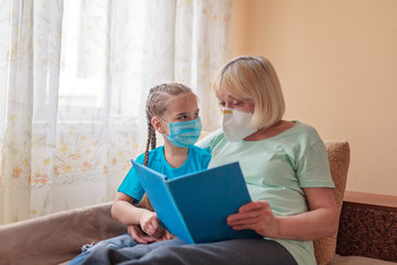 Multigenerational relations, girl and granny wearing respirators lying on sofa and reading a book