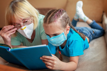 Multigenerational relations, girl and granny wearing respirators lying on sofa and reading a book