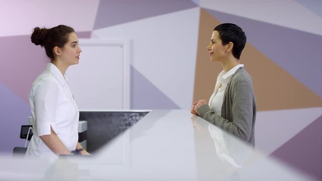Tracking of mature woman smiling and walking up to reception area of modern hospital and taking clipboard from cheerful medical specialist