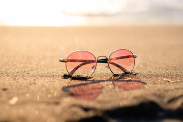 Fototapeta na wymiar Pink sunglasses lie on a sandy beach against the backdrop of the sea at sunset. summer holidays, space for text