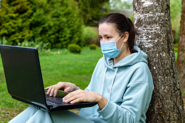 A girl in a tracksuit and a protective mask is sitting in nature, leaning against a birch tree, and holding a laptop on her lap