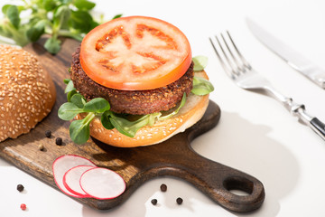 selective focus of delicious vegan burger with radish, tomato and microgreens on wooden boar with black pepper near fork and knife on white background