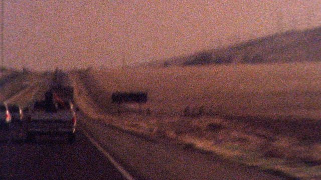 Shaky old looking archival footage drving on 101 highway towards Los Angeles past dessert as sun sets in the distance. Film texture, grit, grain, dust and flash frames create historical look and feel
