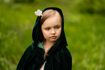 A fairy-tale portrait of a child in the forest, with blond hair, a green velvet Cape with a hood,...