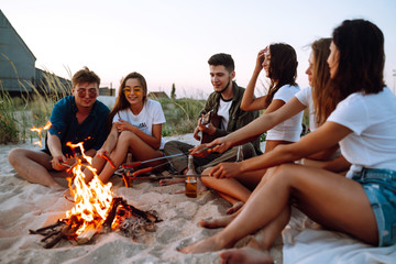Group of young friends sitting on beach and fry sausages. One man is playing guitar. Summer holidays, vacation, relax and lifestyle consept. Camping time.