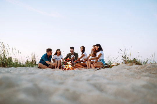 Group of young friends sitting on beach and fry sausages. One man is playing guitar. Summer holidays, vacation, relax and lifestyle consept. Camping time.