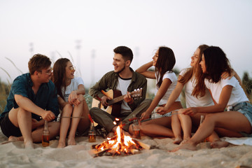 Group of young friends sitting on beach and fry sausages. One man is playing guitar. Summer holidays, vacation, relax and lifestyle consept. Camping time.