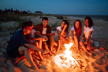 Group of young friends sitting on beach and fry sausages. One man is playing guitar. Summer holidays, vacation, relax and lifestyle consept. Camping time.