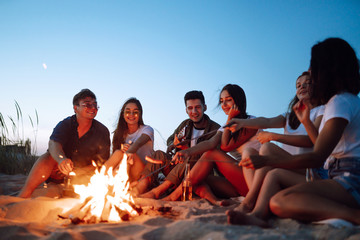 Group of young friends sitting on beach and fry sausages. One man is playing guitar. Summer holidays, vacation, relax and lifestyle consept. Camping time.