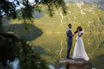 The groom reads the oath of allegiance to the bride standing on a large stone in the lake against the backdrop of the mountains. The bride is crying. Wedding concept. Copy space