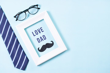 Happy fathers day concept. Top view of tie, mustache, glasses and white picture frame with LOVE DAD text on bright blue pastel background. Flat lay.
