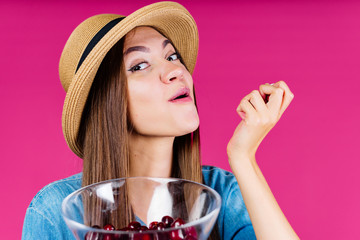 perky and amazed smiling girl in a hat eats cherry berries from a vase in her hand background pink