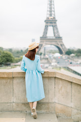 Obraz premium Pretty young caucasian girl walking near stairs, Eiffel Tower in background, Paris. Charming lady in blue dress staying back to camera and looking at the beautiful view of Paris
