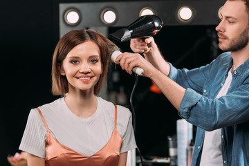 Handsome makeup artist doing hairstyle with hairbrush and hair dryer on smiling model in photo studio