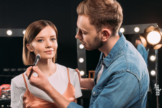 Handsome makeup artist applying face powder on attractive model in photo studio