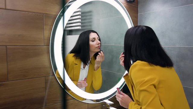 Beautiful confident young business woman in stylish suit putting pomade on her lips while looking into the mirror