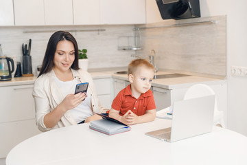 Woman on maternity leave working home online with laptop in kitchen with small child. Concept mom work while in quarantine isolation during Covid-19