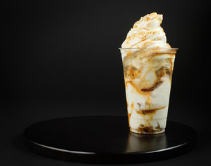 Caramel vanilla ice cream in plastic cup covered with whipped cream candy sprinkles side view on a dark background. Soft vanilla ice cream with caramel topping and chocolate chips.

