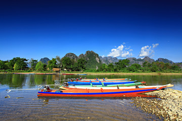 Fototapeta premium Colorful motoboats and few people on the Nam Song River and hotels in Vang Vieng, Vientiane Province, Laos, on a sunny day 