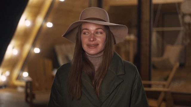  portrait Young Beautiful Woman in hat Walking on the background ethnic triangular wooden house with lanterns at night. Slow motion. Front View. Close up