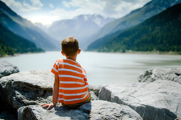 Little boy sitting on rocks with his back looking at the mountain lake.