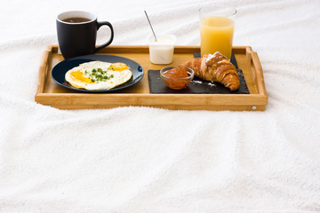 Obraz na płótnie Canvas Wooden coffee tray with fried eggs, croissant with jam, cup of tea, juice and yogurt on white bedding. Breakfast in the bed good morning concept. Romantic Weekend. Vacations. Space for text.