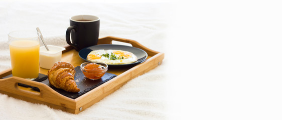 Wooden coffee tray with fried eggs, croissant with jam, cup of tea, juice and yogurt on white bedding. Breakfast in the bed good morning concept. Romantic Weekend. Vacations.Banner. Space for text