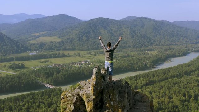 Young man standing victoriously on top of the mountain at sunrise. Aerial view.