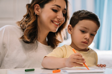 cheerful mother looking at adorable son drawing with felt-tip pen