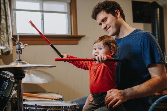 Father Teaching Baby Boy To Play Drums. Parent With Toddler Child Having Fun Spending Time Together. Dad And Kid Playing Music. Family Hobby Activity And Leisure Time During Self Isolation.