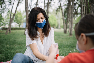 Woman and child sitting in nature and using hand sanitizer. Back of kid holding disinfectant over woman's hands while sitting in park.