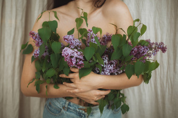 Beautiful girl covering naked upper body with blooming lilac flowers. Creative image of tenderness and sensuality. Young woman with growing from denim jeans lilac branches, cropped view.