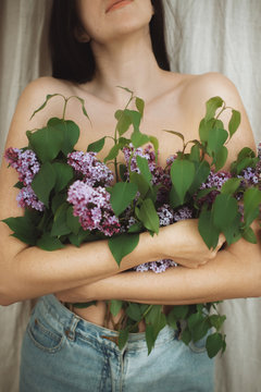 Beautiful girl covering naked upper body with blooming lilac flowers. Creative image of tenderness and sensuality. Young woman with growing from denim jeans lilac branches, cropped view.