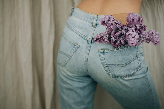 Woman posing with lilac flowers in denim jeans pocket on rustic background. Blooming lilac flowers  in back pocket. Creative image. Sensual atmospheric mood. Hello spring