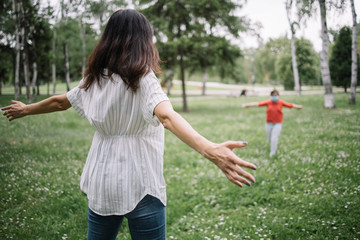 Back view of mother with opened arms waiting her child for a hug. Back of woman with spread hands looking at running little girl in park.