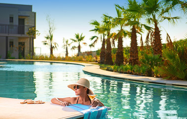 Obraz premium Young woman with martini glass, hat, bikini in blue water swimming pool at sunset hotel party