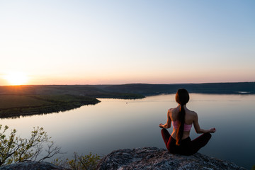 Woman doing yoga and meditating in lotus position on the background of sky, river and sunset