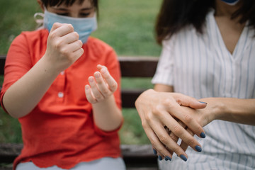 Mother and daughter spreading hand sanitizer on their hands outdoor. Close-up view of cropped woman and little girl sanitizing their hands with disinfectant while sitting on bench.