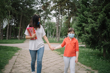 Lady and child holding their hands while walking in park. Mother holding her little daughter's hand while walking on park alley and wearing antivirus masks.