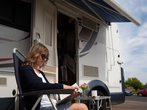A lady sits outside her recreational vehicle reading a book shaded by an awning.She is parked on a drive with cars and street in background.Stay at Home.Image