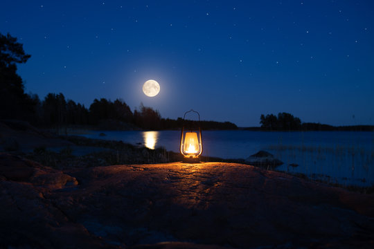 Vintage lantern in forest on shore of lake. Oil lamp with warm orange light on the rock. Beautiful moon on the sky.