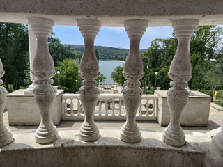 Fototapeta premium Stone stairs to the lake. Around the park with green trees and firs. Blue sky with clouds. View through the white columns.