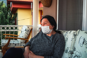 Middle aged caucasian woman with medical face mask, resting on cpuch on veranda during COVID-19 threat. Woman with mask on her terrace is resting, sleeping, looking, talking.