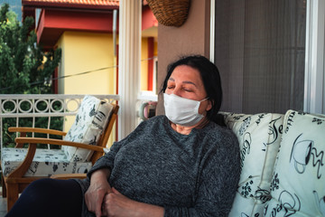 Middle aged caucasian woman with medical face mask, resting on cpuch on veranda during COVID-19 threat. Woman with mask on her terrace is resting, sleeping, looking, talking.