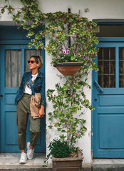 Pretty fashionable woman standing in doorway of old house. Cool dressed lady in front of turquoise wooden door with flowers around.