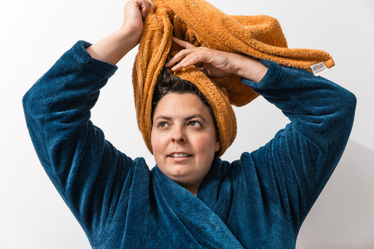 es una mujer.  de 40 años y sobrepeso caucásica con lleva una toalla marrón naranja enrollada en la cabeza para secarse el pelo mojado, lo está enrollando con las manos. lleva un albornoz azul.