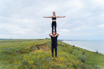 Strong, muscular athlete holding on his shoulders a fit, beautiful sportswoman girl. Acrobats warming up before training, standing on cliff hill on the background of the sea. Tricks akroyoga in pairs