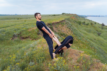 Naklejka na ściany i meble Young caucasian couple practicing acro yoga together on the hill near the sea. Couple, partner yoga. Athlete flexing beautifully and holding girl with his trained hands. Sportswoman stretching