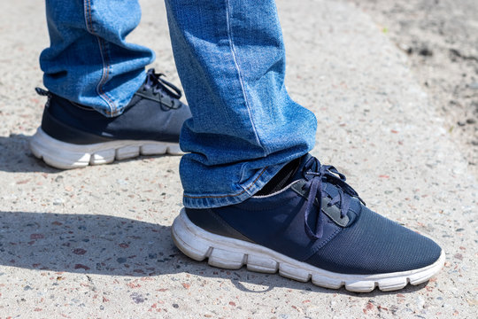 Men's legs in blue jeans and sneakers with white soles stand on the pavement on a sunny day