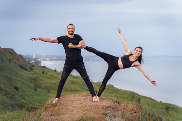 Athletic, pumped up, muscular, beautiful couple in love posing on the edge of the hill. Doing stretching and pair acro yoga on the background of the sea in the sunset. Girl leaned towards the cliff